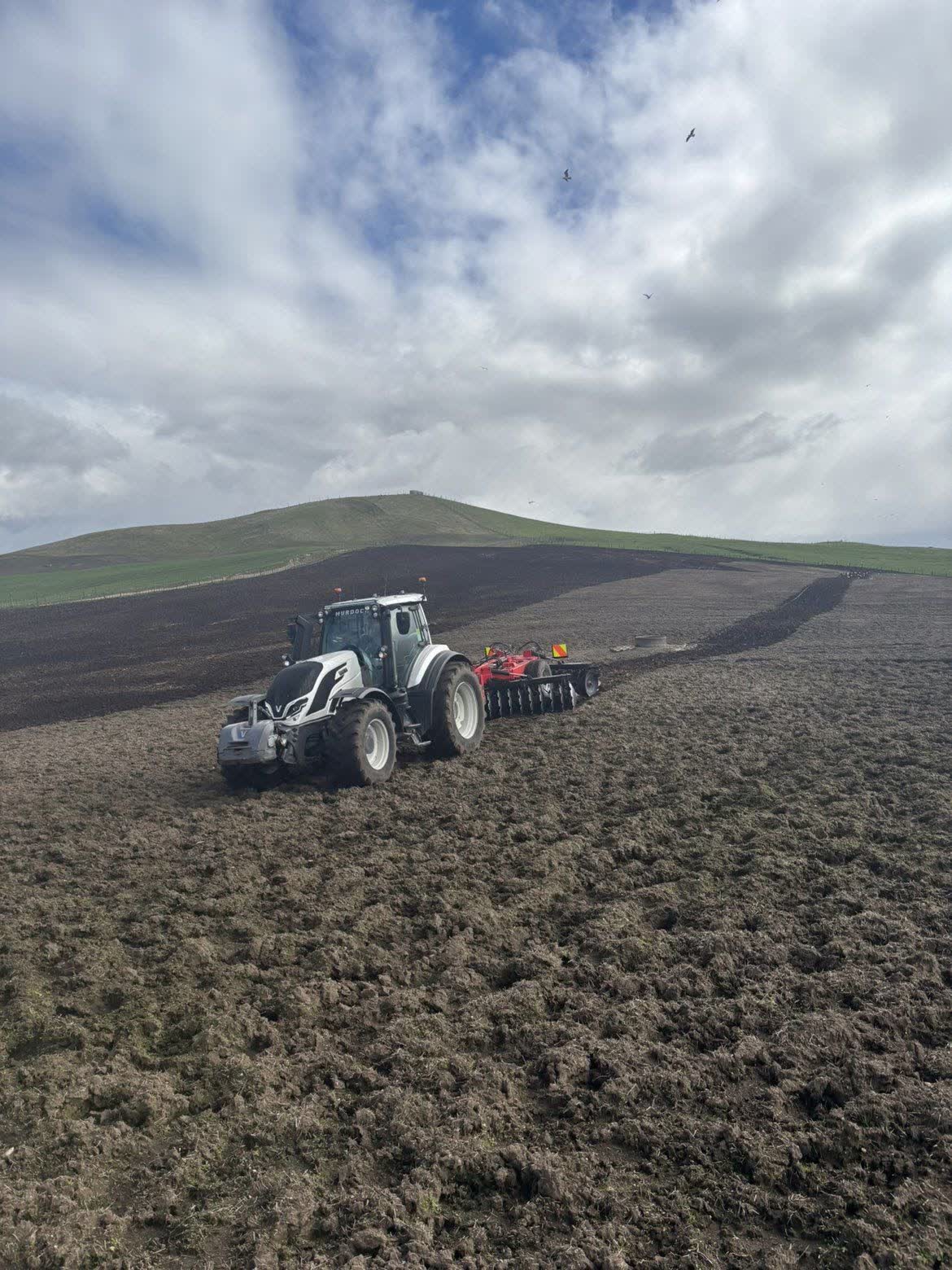 Tractor and implement working across a cultivated field with low hills beyond.